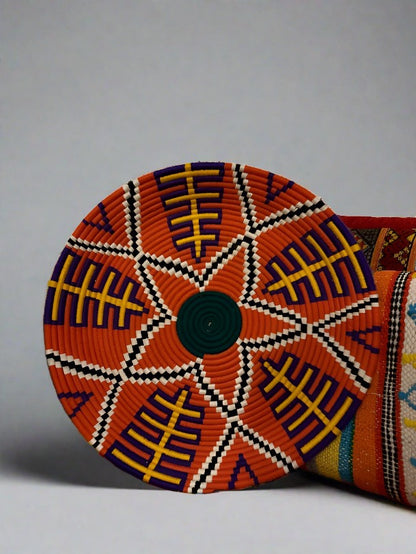 A large handmade decorative plate with a geometric and colorful pattern, featuring a central star-like design surrounded by diamond-shaped patterns in a spiral pattern, with borders in a contrasting color.